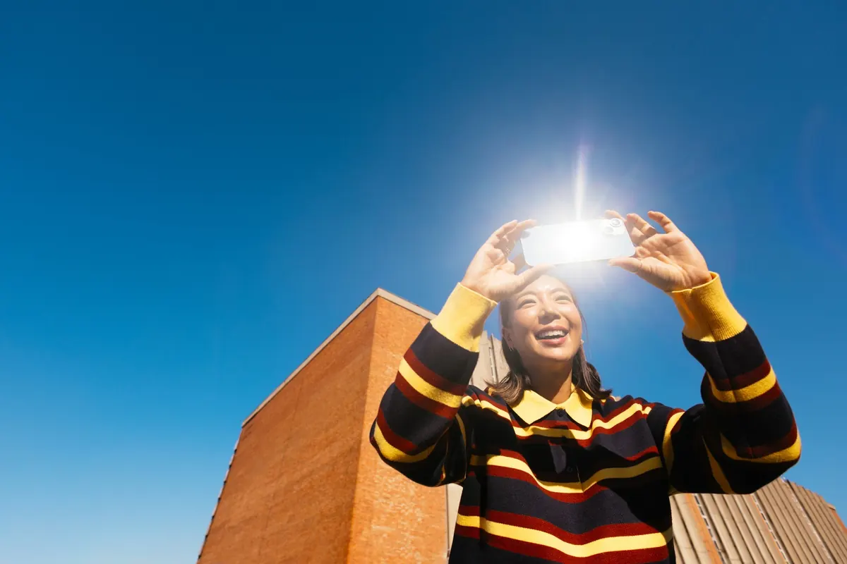 A young woman holding a phone up to the sun against a blue sky